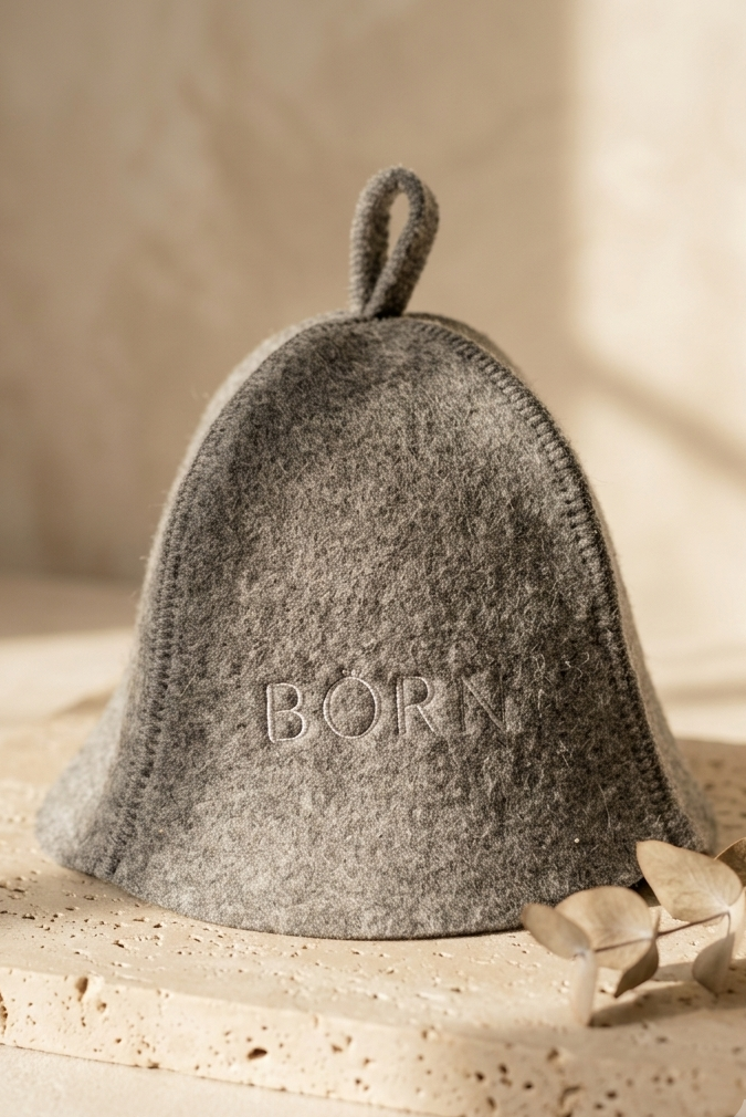 A grey felt hat with a loop at the top and the word 'BORN' embroidered on the front, displayed on a textured beige surface with dried leaves in the foreground.